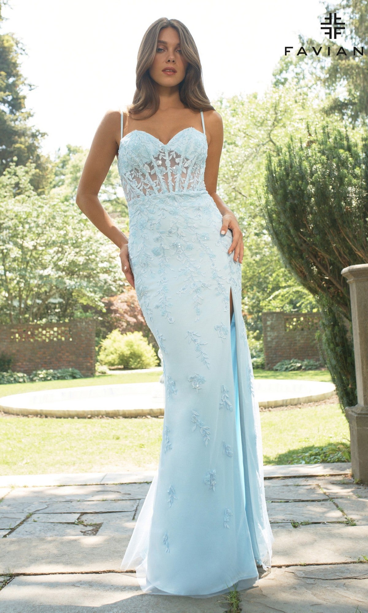 A woman stands in a garden wearing the Faviana 11439 long prom dress, featuring light blue lace embroidery, a sheer corset bodice, thin straps, and a thigh-high slit.
